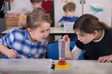 The picture shows two girls enthusiastically working on an experiment with water. They are examining a transparent container of liquid, while other children in the background are also busy with experimental materials.