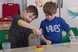 The image shows two children enthusiastically conducting an experiment with a transparent container containing water and possibly something else. They appear to be intensely observing and interacting with each other while conducting the experiment in the educational setting.