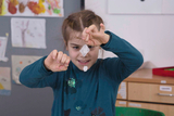 The picture shows a girl enthusiastically conducting an experiment. She is holding a pendant with a crystal structure in her hand while standing in a creative environment with drawings and craft materials.