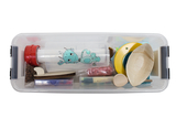 The picture shows a transparent storage box filled with various experimental materials, including a container with a cartoonish blue creature, small wooden parts, plastic parts and other creative utensils. These materials are ideal for practical educational projects in the German school system.