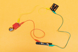The picture shows a simple electrical circuit with a battery, wires and a small bell detector. The colored wires are connected to transmit the signal to the bell and illustrate basic principles of electricity.
