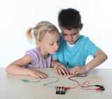 The picture shows two children working intently on an experiment with batteries and wires. They use materials to create a circuit, providing a fun introduction to the basics of electricity.
