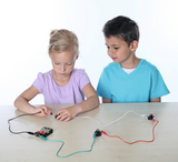 The picture shows two children working on an experiment with electrical circuits. The girl is connecting cables to a battery and a lamp while the boy looks on with interest.
