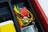 The picture shows a collection of experimental materials in a red storage box. You can see various cables with crocodile clips, a small battery and other electrical components that are used for experimental purposes.