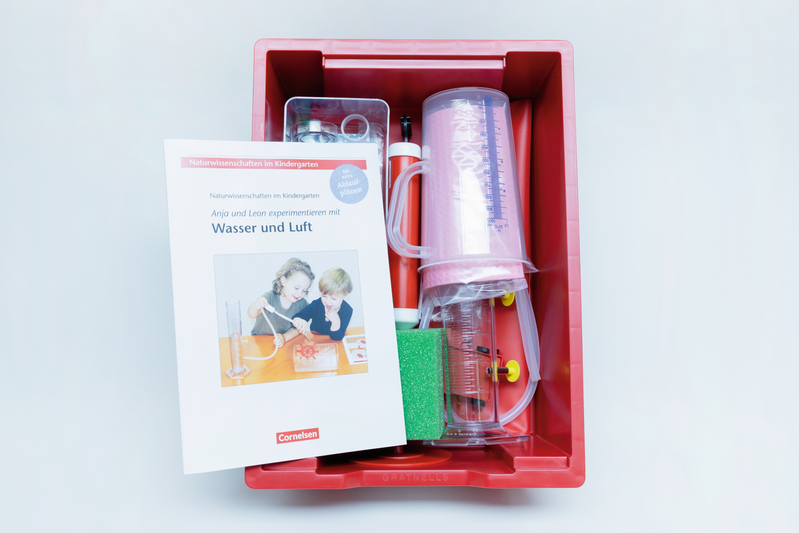 The picture shows a red box containing experimental materials for children, including measuring cups, tubes and an information booklet. The booklet is entitled &quot;Anja and Leon experiment with water and air&quot; and is intended for use in kindergarten.