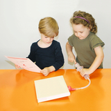 The picture shows two children sitting at an orange table. One child is holding a sheet of paper while the other is working with an experimental device attached to an open book.