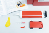 The picture shows experimental materials, including a red air pump, a yellow balloon, a small red plastic vehicle and accessories. In the background you can see an open book that provides instructions for experiments with water and air.