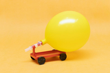 The picture shows a yellow balloon model mounted on a small red cart. The cart has four black wheels and is probably used as an experimental device to illustrate physical principles such as air pressure and motion.