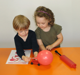 The picture shows two children working together on an experiment with a red balloon and a small cart. There is a worksheet on the table and an air pump next to the children.