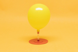 The image shows a yellow balloon standing on a red base and connected by a transparent tube. The whole composition has a cheerful, inviting look against a solid yellow background.