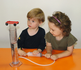 The picture shows two children working on an experimental device. One child is holding a syringe while the other is pointing to a cylinder filled with water and with a small vehicle on top.