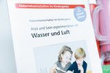 The picture shows a workbook entitled &quot;Natural Science in Kindergarten&quot;, which focuses on experiments with water and air. It shows two children, Anja and Leon, working together on an experiment.