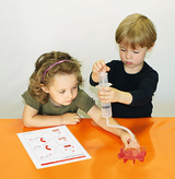 The picture shows two children working on an experiment. The girl is following instructions on a piece of paper, while the boy is using a syringe to inject a liquid into a device.