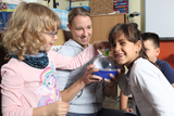 The picture shows a teacher conducting an experiment with two children. The children are excited and interacting with a scientific device while another child watches attentively in the background.