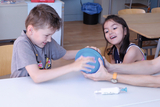 The picture shows a boy and a girl working together on a blue ball. They appear to be doing the task with enthusiasm and joy, while an adult&#39;s hand supports them.