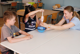 The picture shows two children and a teacher working together on an experiment in the classroom. The girl is operating an apparatus while the teacher intervenes to assist, and the boy is watching the activity attentively.