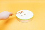 The image shows a hand using an orange mallet to hit a white, round disk on a yellow background. There are several small screws on the disk.