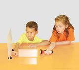 The picture shows two children experimenting on an orange table. The girl is holding a flashlight while the boy is pointing to a transparent box and a reflective surface, suggesting an exciting learning experiment.