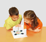The picture shows two children, a boy in a yellow T-shirt and a girl in an orange T-shirt, curiously looking at an experimental project. In front of them is a surface with mirrors and various objects that invite experimentation.