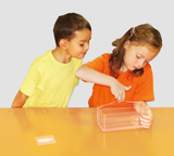 The picture shows two children working at a table with a transparent container. The girl in the orange shirt is examining the container while the boy in the yellow shirt is watching with interest.
