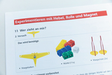 The picture shows instructions for an experiment that involves levers, pulleys and magnets. It lists the materials needed, including a balance beam, various colored cubes and two weighing pans.