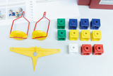 The picture shows various experimental learning toys, including colorful cubes in different colors and shapes and two red holders with yellow elements. In the background, work materials and instructions for experimentation in the educational field are visible.