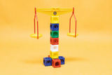 The picture shows a colorful seesaw or balance made of various plastic cubes supported by a yellow frame with red hanging bowls. The construction is set against an orange background and is suitable for educational experiments.