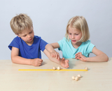The picture shows two children sitting at a table playing with wooden beads and a yellow ruler. They appear to be conducting an experiment together, with one of the children playing with the beads while the other looks closely.