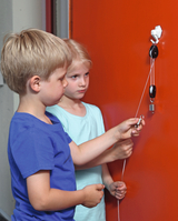 The picture shows two children working on an experiment with a suspension and a string. The orange wall in the background creates a vivid contrast to the children&#39;s activities, who appear curious and focused.
