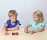 The picture shows two children sitting at a table experimenting with toy vehicles. The girl in the blue t-shirt is smiling while the boy is staring intently at his red vehicle.