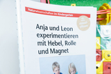 The picture shows a page from a book entitled &quot;Natural Sciences in Kindergarten&quot; in which children named Anja and Leon are shown experimenting with a lever, pulley and magnet. In the background, various experimental materials intended for educational purposes are visible.