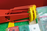 The image shows various educational experiment materials, including a red and yellow device and a selection of colored plates. These materials are arranged on a green base and appear to be designed for hands-on learning activities.