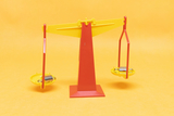 The picture shows a colorful red and yellow seesaw with two bowls balancing on a central support. This seesaw is ideal for experiments on weight and mass distribution in educational settings.
