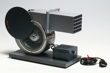 The picture shows an experimental device in the form of a fan with a round lid and a rectangular outlet. The device is operated with a power cable and is suitable for educational purposes in the field of physics or technology.