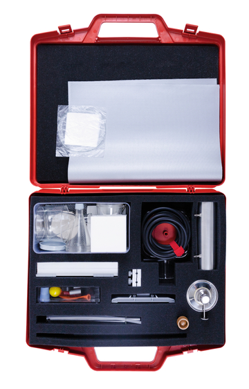 The image shows a red suitcase containing a variety of experimental materials and equipment, including measuring cups, tubing, and a heat source. The careful arrangement of the utensils suggests practical use in the educational field.