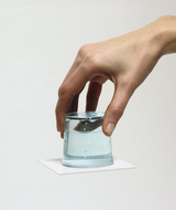 The image shows a hand lifting a glass of water that is placed on a white sheet of paper. The scene conveys an experimental character, possibly for demonstrations in educational contexts.