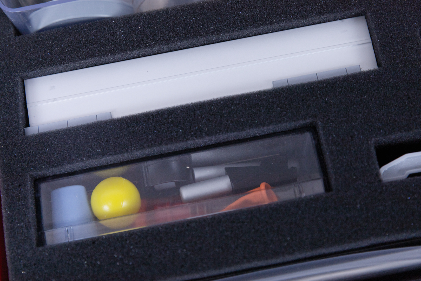 The picture shows a storage box containing various experimental materials. Visible objects include a yellow ball, several small containers and a tool, all well encapsulated in black foam.