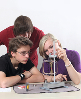 The picture shows three young people working concentratedly on an experimental setup. In front of them is an apparatus with a transparent chamber while they examine various materials and tools on a table.