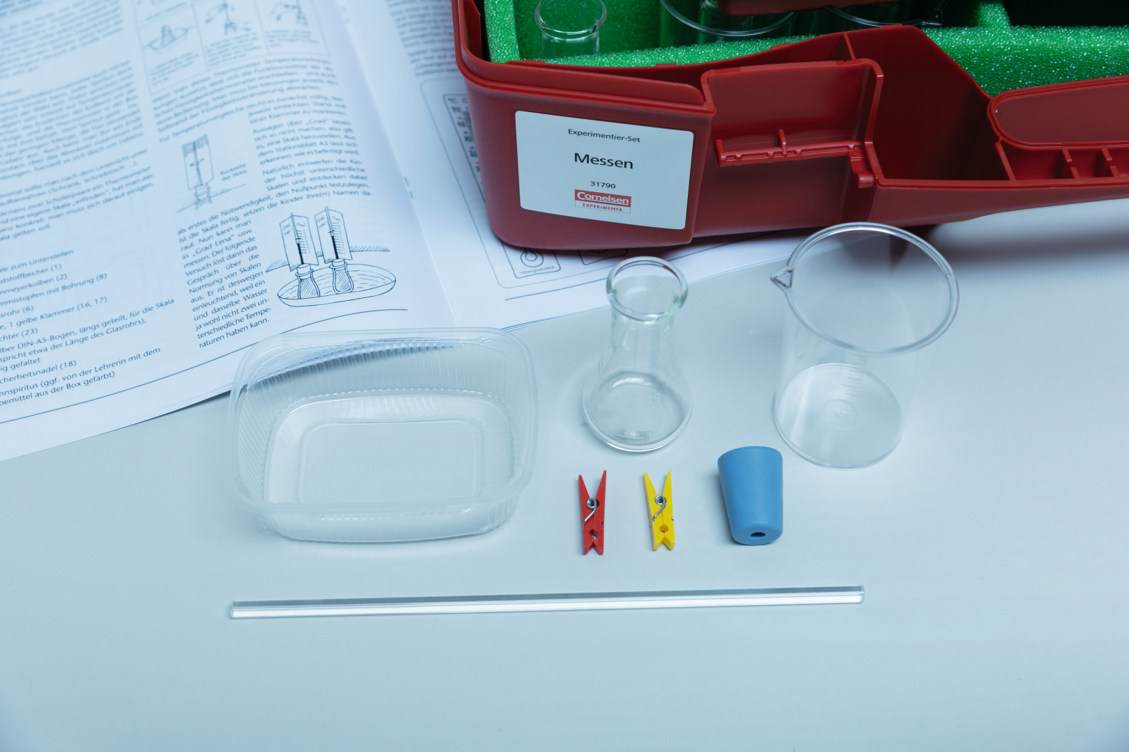 The picture shows an experiment set with various glass containers, a measuring cylinder, red and yellow clamps, a blue object, a transparent container and a straw. The set is placed on a manual for experiments and promotes learning in the natural sciences.