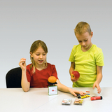 The picture shows two children at a table experimenting with different objects. One child is holding an apple and the other an orange on a scale while they examine other materials such as pens and decks of cards.