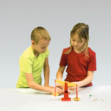 The picture shows two children experimenting together on a table scale. Various small objects are lying on the table and the children appear to be comparing the weights.