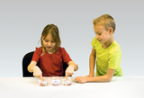 The picture shows two children standing at a table dipping their fingers into three glasses of liquid. The glasses are labeled and appear to be part of an experiment investigating various substances.
