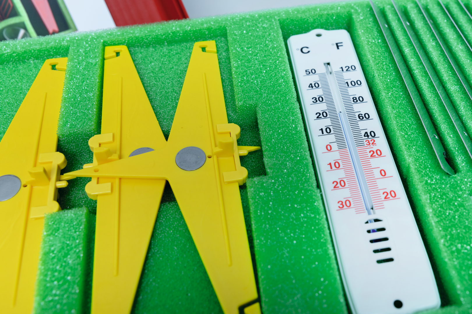 The image shows a carefully arranged collection of experiment tools, including yellow cutters and a thermometer, resting on a green foam pad. These materials are designed for use in education and encourage hands-on learning experiments.