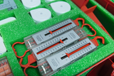 The picture shows two precision scales with orange handles, which are placed in a green foam insert. In the background you can see other experimental equipment and accessories that are suitable for educational purposes.