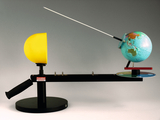 The image shows an experimental device that connects half a yellow dome and a globe with a pointer. It is used to illustrate physical principles, possibly related to light and shadow and the rotation of the earth.