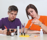 The picture shows two young people taking part in an experiment. The young woman in an orange T-shirt is holding a test tube while the boy in a purple T-shirt is watching attentively; there are various containers and measuring devices on the table.