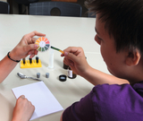 The picture shows two students conducting an experiment by dipping a test strip into a solution. In the background you can see various test tubes and a cover sheet for notes.