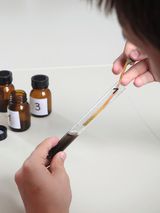 The image shows a hand holding a test tube sample while a strip is dipped into the liquid. In the background are several brown vials with numbers indicating different chemicals or solutions.