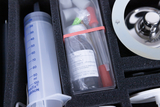 The picture shows a box with various experimental materials, including a syringe and test tubes. The items are stored in a foam-padded interior, which allows the utensils to be transported safely.