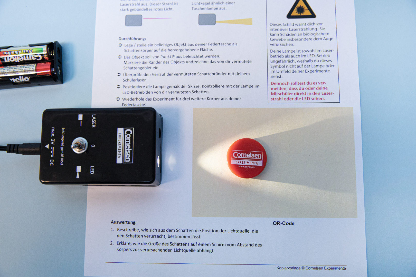 The picture shows an experiment set with a laser device and instructions describing how to carry out an experiment on shadow formation. On a white sheet of paper there is a red toy or token on which the light from the laser is projected.